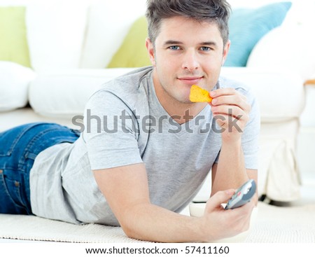 Relaxed young man eating crisps holding a remote lying on the floor looking at the camera