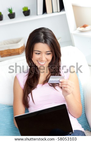Delighted asian woman using her laptop holding a card sitting on a sofa at home
