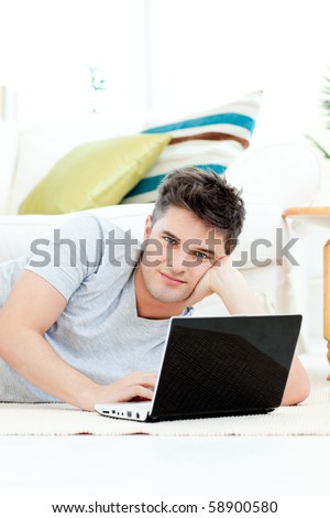 Charismatic young man using his laptop lying on the floor at home looking at the camera