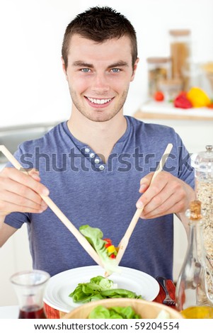 Handsome young man eating a salad smiling at the camera in the kitchen