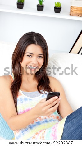 Glowing asian woman holding a cellphone smiling at the camera sitting on a couch at home