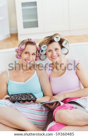 Smiling female friends with hair rollers eating chocolate reading a magazine at home on a sofa