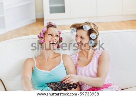 Laughing female friends with hair rollers eating chocolate at home on a sofa