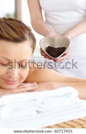 Bright woman lying on a massage table in a spa center with other woman holding a bowl in the shape of a heart with chocolate