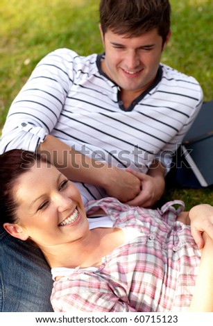 Joyful couple of students sitting on grass and smiling at the ca