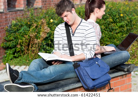 Couple of students using a laptop and reading a book sitting in the campus of their university