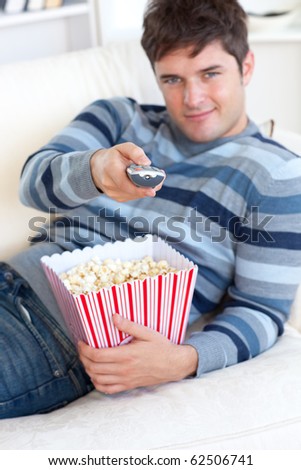 Relaxed young man eating popcorn and holding a remote lying on the sofa at home