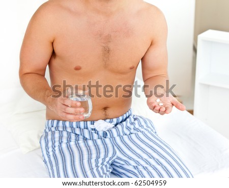 Depressed young man sitting on his bed and holding a glass of water and pills in his bedroom at home