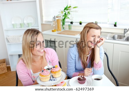 Two cheerful female friends eating pastries and drinking coffee in the kitchen at home