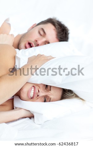 Scenic in bedroom of loving young couple lying in bed and holding pillow on the ears because of snoring boyfriend.