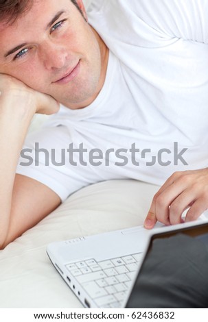 Handsome man writing on his laptop lying in his bedroom smiling at the camera