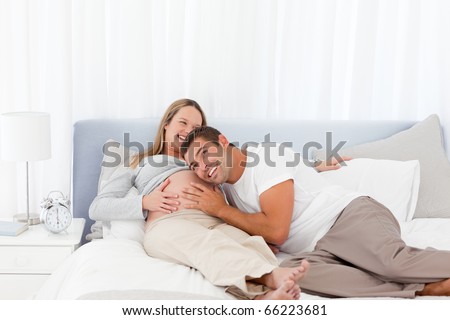 Funny man putting his head on the belly of his pregnant wife while relaxing on the bed