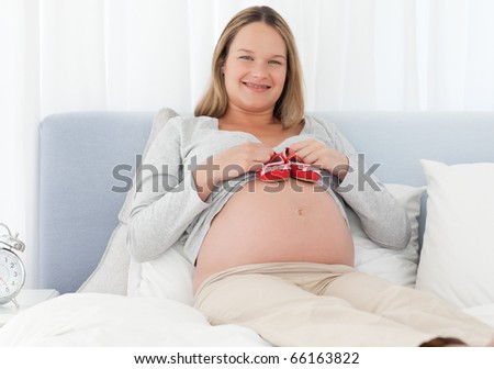 Cheerful pregnant woman putting baby shoes on her belly on a bed