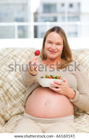 Future mother showing a strawberry to the camera sitting on the sofa