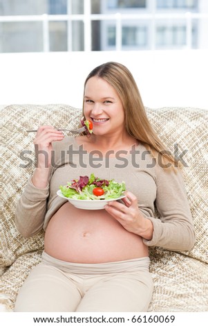 Happy pregnant woman enjoying a salad sitting on the sofa at home