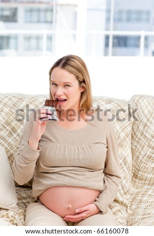 Pretty future mom eating a chocolate bar sitting on the sofa at home
