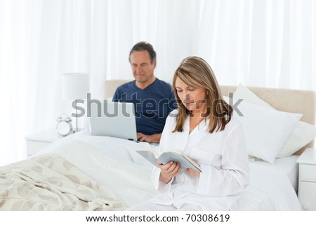 Senior looking at his laptop while her wife is reading at home