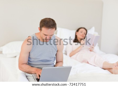 Man working on his laptop while his wife is reading a book on the bed