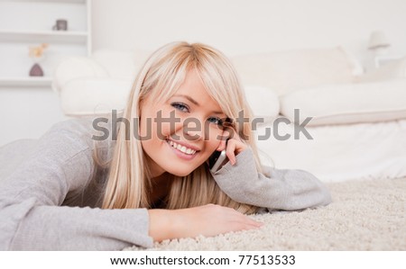 Attractive happy blond woman talking on cell phone lying down on a carpet in the living room