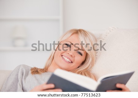 Smiling blonde woman reading a book while lying on a sofa