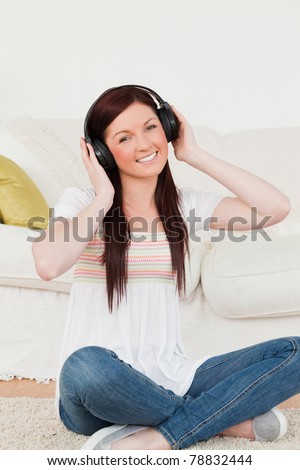 Good looking red-haired woman listening to music with headphones while sitting on a carpet in the living room