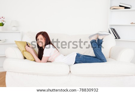 Good looking red-haired female writing a text on her phone while lying on a sofa in the living room