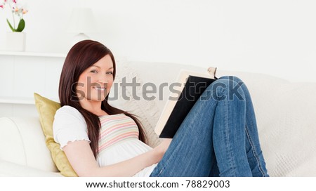 Beautiful red-haired woman reading a book while lying on a sofa in the living room