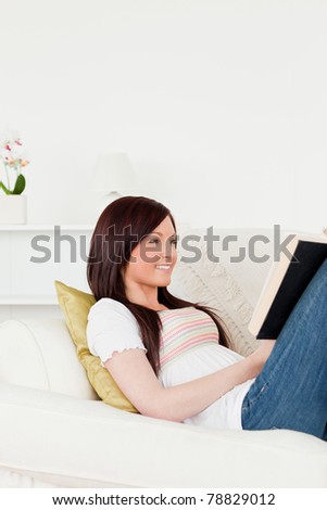 Charming red-haired woman reading a book while lying on a sofa in the living room