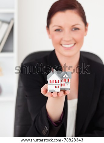 Young gorgeous red-haired woman in suit holding a miniature house while sitting in an office