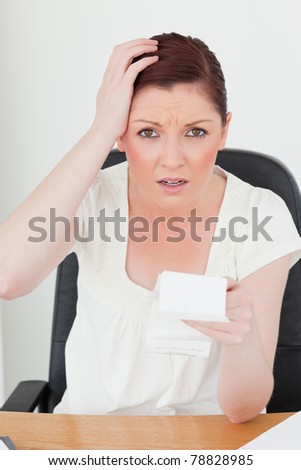 Young attractive red-haired female being scared by the amount of the receipt while sitting at a desk
