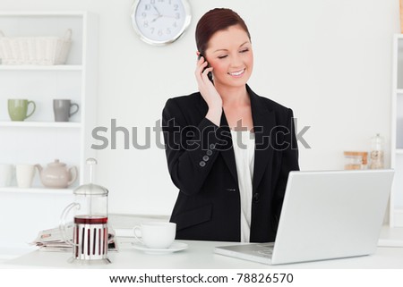 Pretty red-haired woman in suit relaxing with her laptop while phoning in the kitchen in her apartment