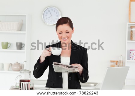 Pretty red-haired woman in suit reading the newspaper while having her breakfast in the kitchen in her apartment