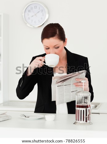 Good looking red-haired woman in suit reading the newspaper while having her breakfast in the kitchen in her apartment