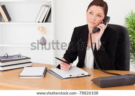 Good looking red-haired woman in suit writing on a notepad and phoning while sitting in an office