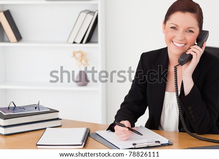 Pretty red-haired woman in suit writing on a notepad and phoning while sitting in an office