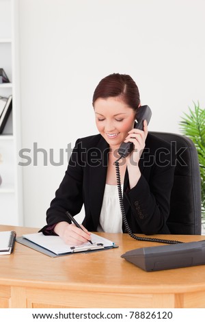 Young attractive red-haired woman in suit writing on a notepad and phoning while sitting in an office