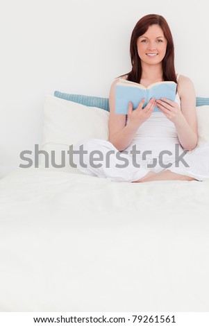 Charming red-haired woman reading a book while sitting on her bed