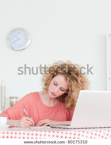 Attractive blonde woman writing while working with her laptop in the kitchen in her apartment
