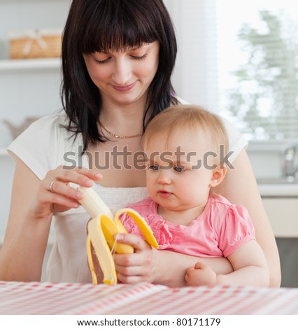 Charming brunette woman pealing a banana while holding her baby on her knees in the kitchen
