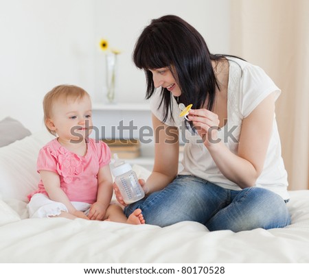 Charming brunette female showing a feeding bottle to her baby while sitting on a bed in her apartment