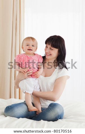 Cute brunette woman holding her baby on her knees while sitting on a bed in her apartment