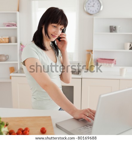 Good looking brunette female on the phone while relaxing with her laptop in the kitchen