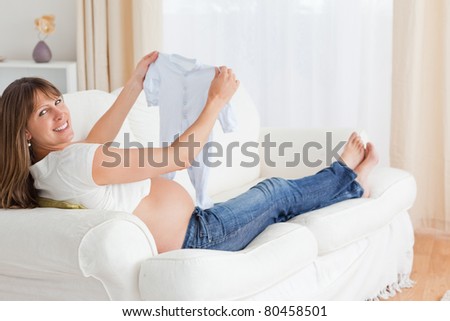 Beautiful pregnant woman holding a baby grow and posing while lying on a sofa in her apartment
