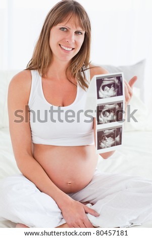 Beautiful pregnant female showing an ultrasound scan while sitting on a bed in her apartment