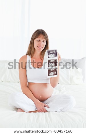 Pretty pregnant female showing an ultrasound scan while sitting on a bed in her apartment