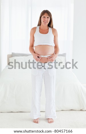 Beautiful pregnant female using a tape measure while standing in her bedroom