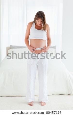 Attractive pregnant female using a tape measure while standing in her bedroom