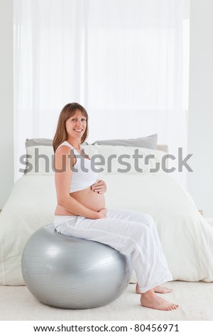Beautiful pregnant female caressing her belly while sitting on a gym ball in her bedroom