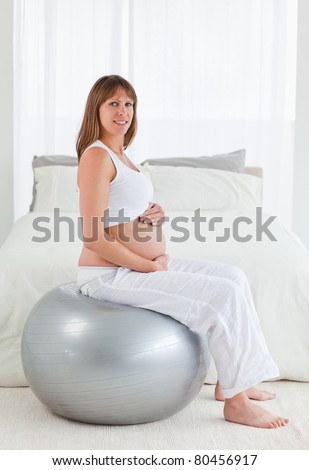 Attractive pregnant female caressing her belly while sitting on a gym ball in her bedroom