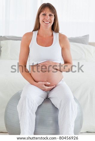 Charming pregnant female caressing her belly while sitting on a gym ball in her bedroom